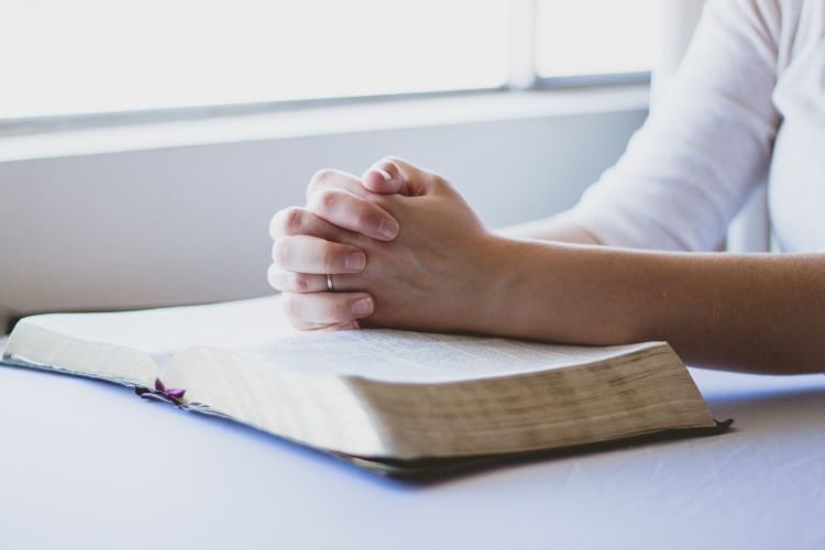 An open Bible on a table with a woman's hands folded in prayer on top of it